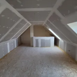 Poddasze w Bartoszycach w trakcie adaptacji: widoczne płyty gipsowo-kartonowe na ścianach i suficie, częściowo zaszpachlowane, oraz surowa podłoga z płyt OSB. W tle okno dachowe.