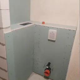 Montaż instalacji podtynkowej WC na ścianie z płyty kartonowo-gipsowej w trakcie remontu łazienki, widoczne elementy mocujące i rury kanalizacyjne.