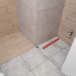 Układanie płytek w łazience: widok na narożnik z ułożonymi płytkami na ścianie imitującymi drewno i szare płytki podłogowe, z użyciem krzyżyków dystansowych, poziomicy i gumowego młotka.