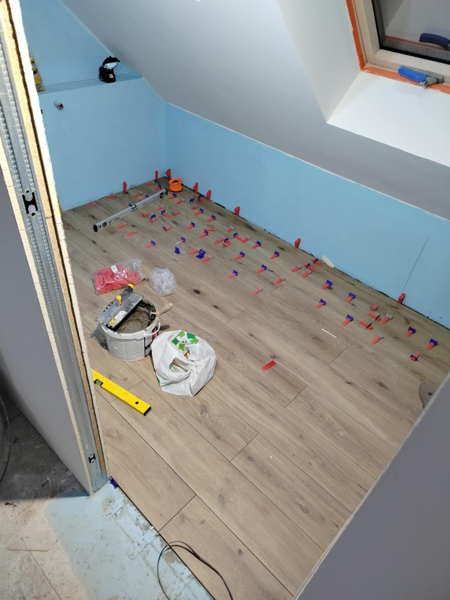 Układanie paneli podłogowych w pokoju na poddaszu. Widoczne narzędzia, poziomica i niebieskie klipsy dystansowe. Ściany pomalowane na jasny niebieski kolor.