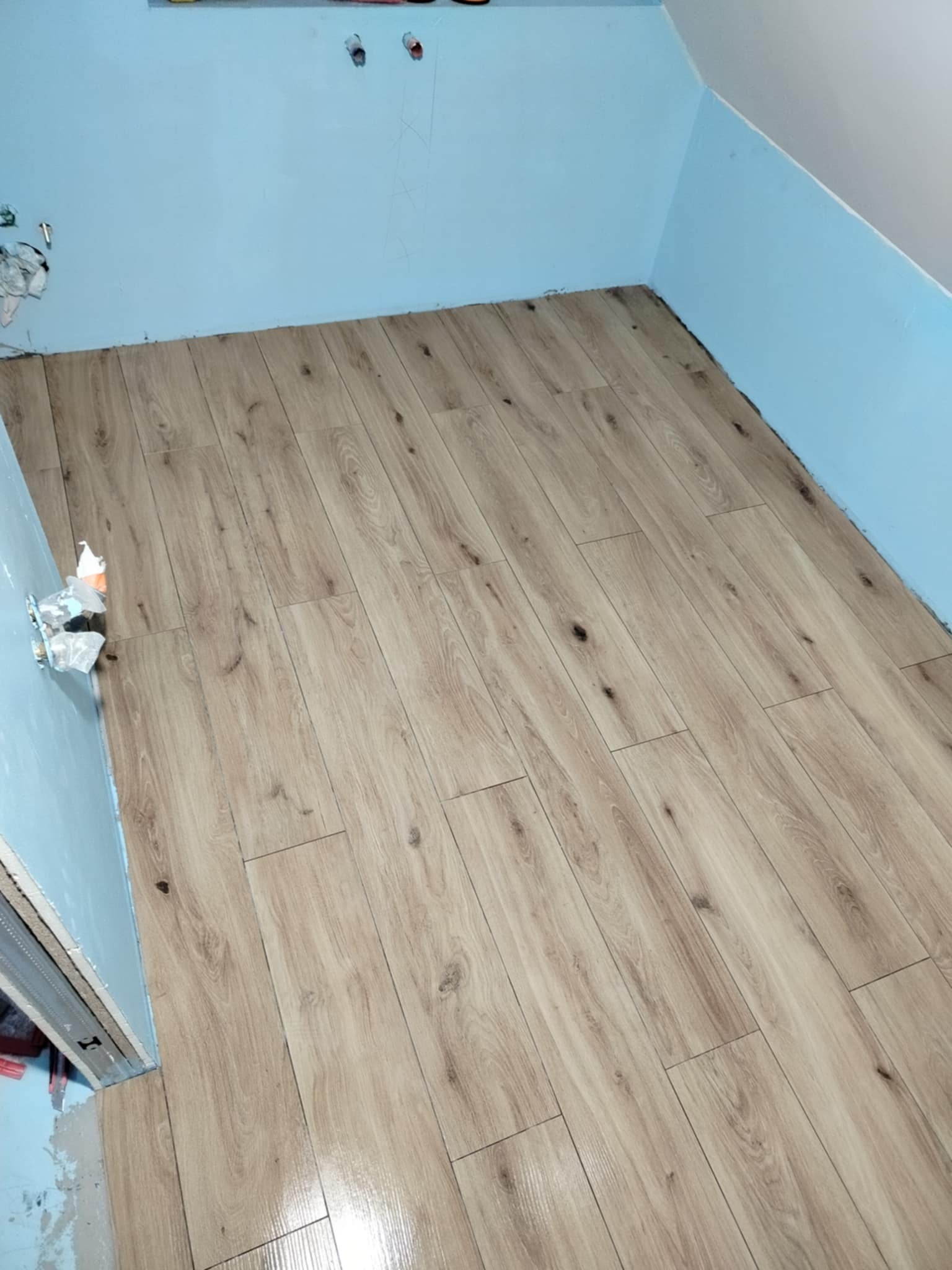 Ukończona podłoga z paneli drewnopodobnych w pokoju z niebieskimi ścianami. Widoczne niedokończone wykończenia przy ścianach i instalacji.