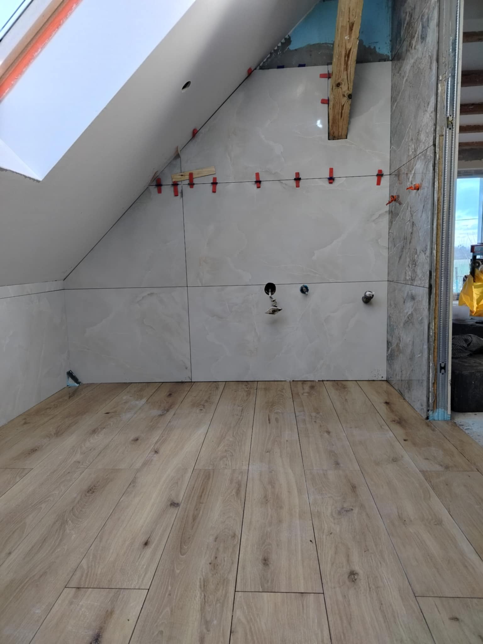 Wykończenie łazienki na poddaszu: ułożone płytki imitujące marmur z poziomowaniem, drewnopodobna podłoga, okno dachowe. Widoczne elementy instalacji.