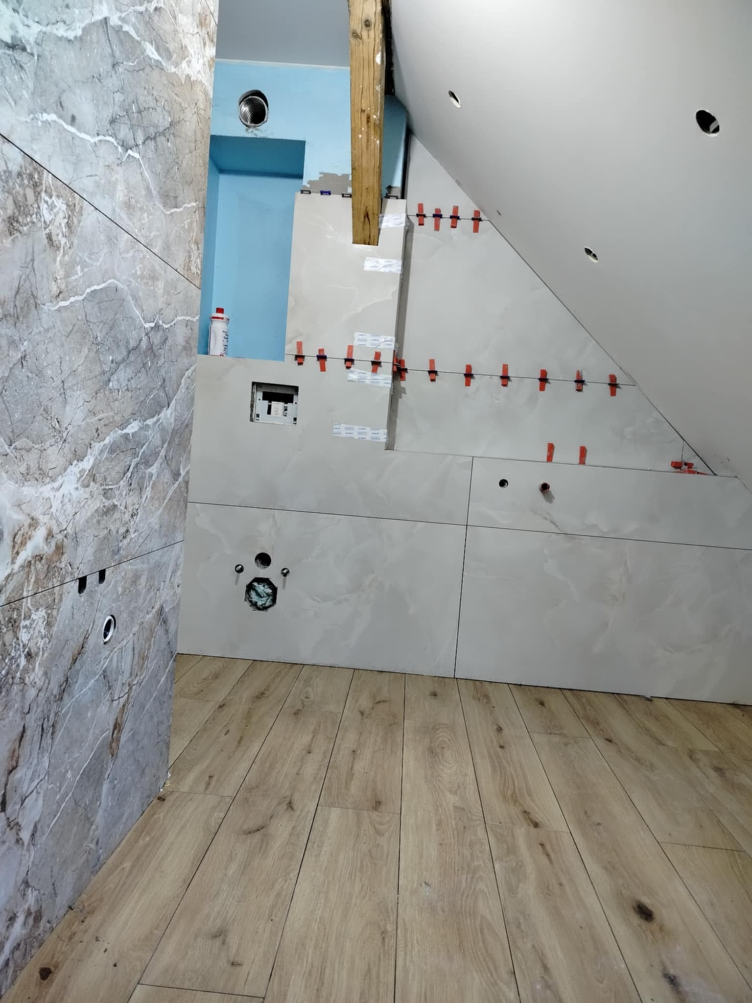 Układanie płytek w łazience na poddaszu. Widoczne płytki imitujące marmur na ścianie i podłodze, oraz białe płytki na skośnej ścianie z czerwonymi krzyżykami dystansowymi.