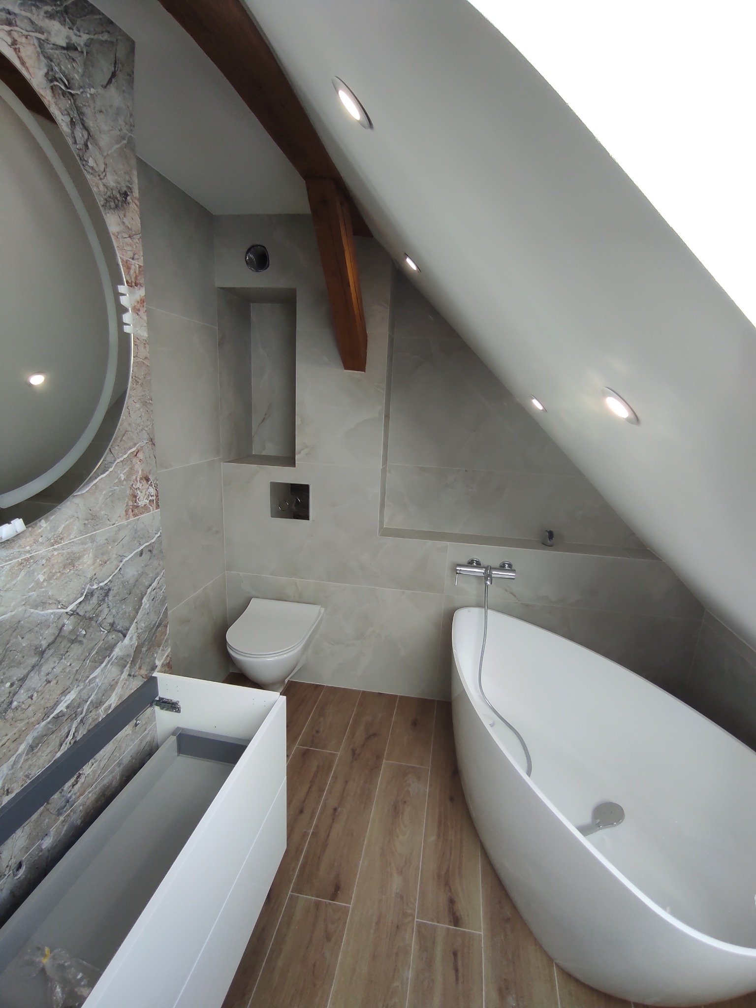 Nowoczesna łazienka na poddaszu z wolnostojącą wanną, marmurową ścianą z okrągłym lustrem i drewnianą belką stropową. Umywalka z szafką i toaleta w neutralnej kolorystyce.