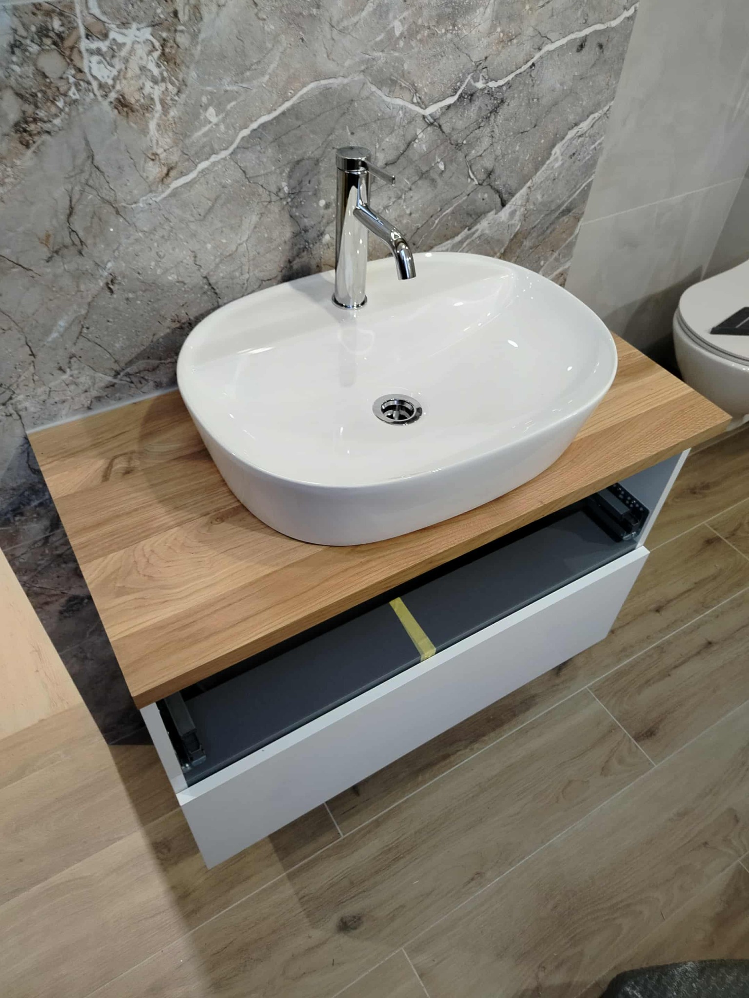 Nowoczesna łazienka: biała, owalna umywalka nablatowa z chromowaną baterią na drewnianym blacie, szara marmurowa ściana i drewnopodobna podłoga.