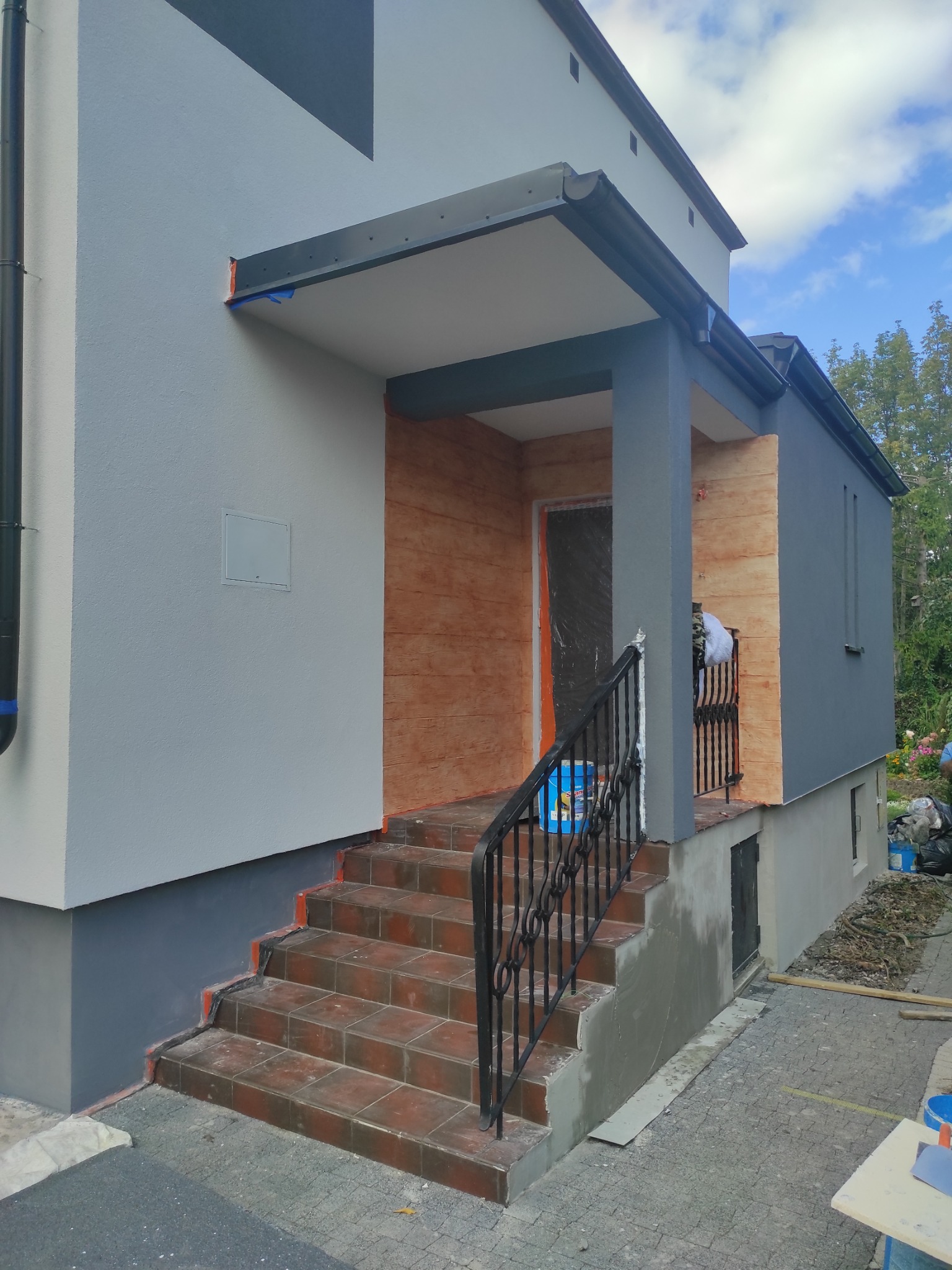 Dwukolorowa elewacja domu z wejściem, schodami z terakoty i balustradą. Część elewacji imituje drewno. Widoczne ślady prac wykończeniowych.