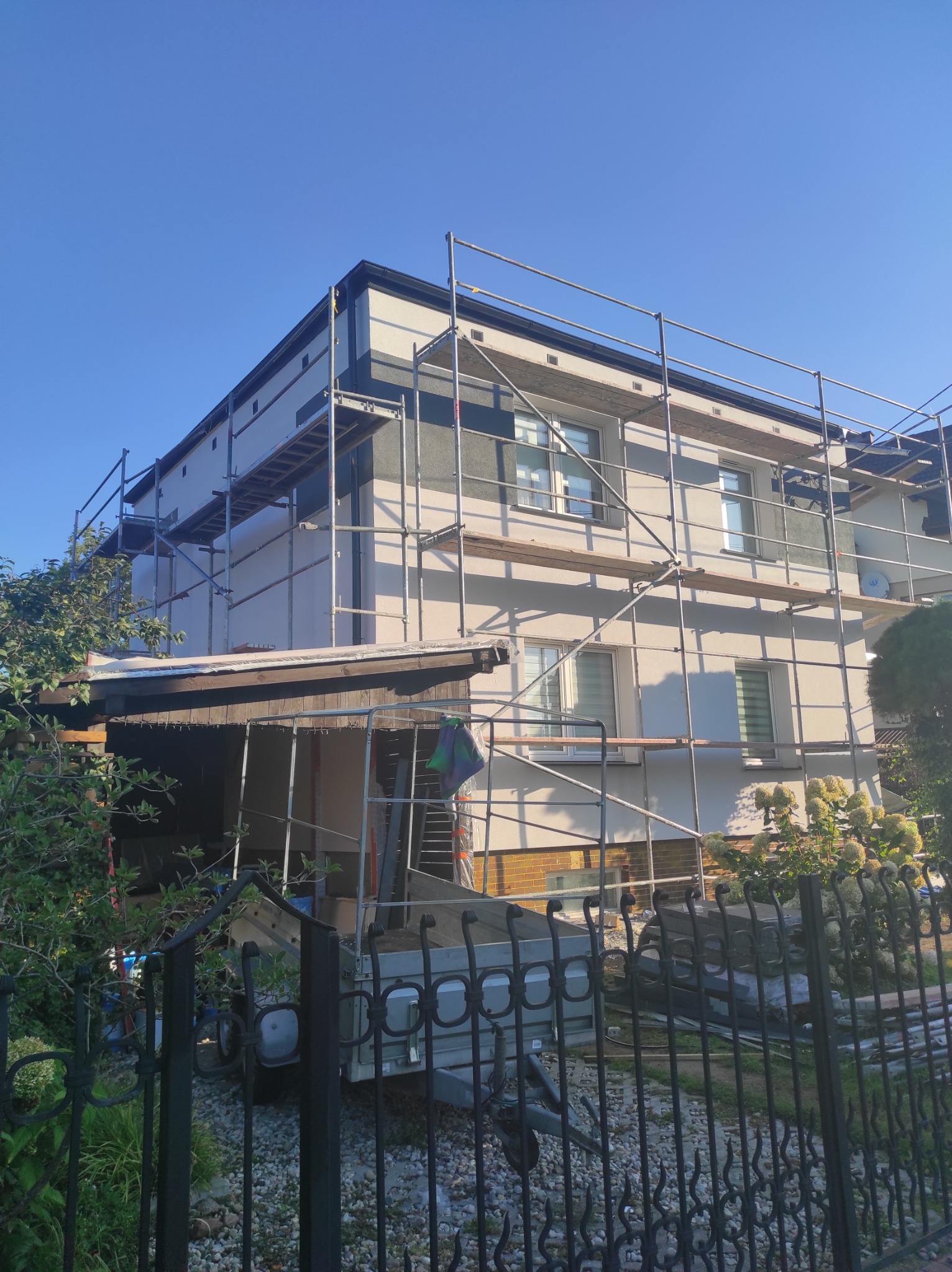 Dwupiętrowy dom w trakcie prac elewacyjnych, otoczony rusztowaniami. Część ściany pomalowana na szaro, reszta na biało. Widoczny fragment przyczepy i ogrodzenia.