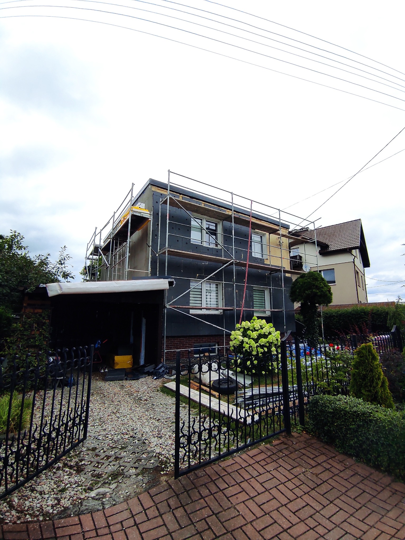 Dwupiętrowy dom w trakcie ocieplania, z widocznymi rusztowaniami i czarną izolacją termiczną. Część elewacji jeszcze nieocieplona. Otwarta brama wjazdowa.