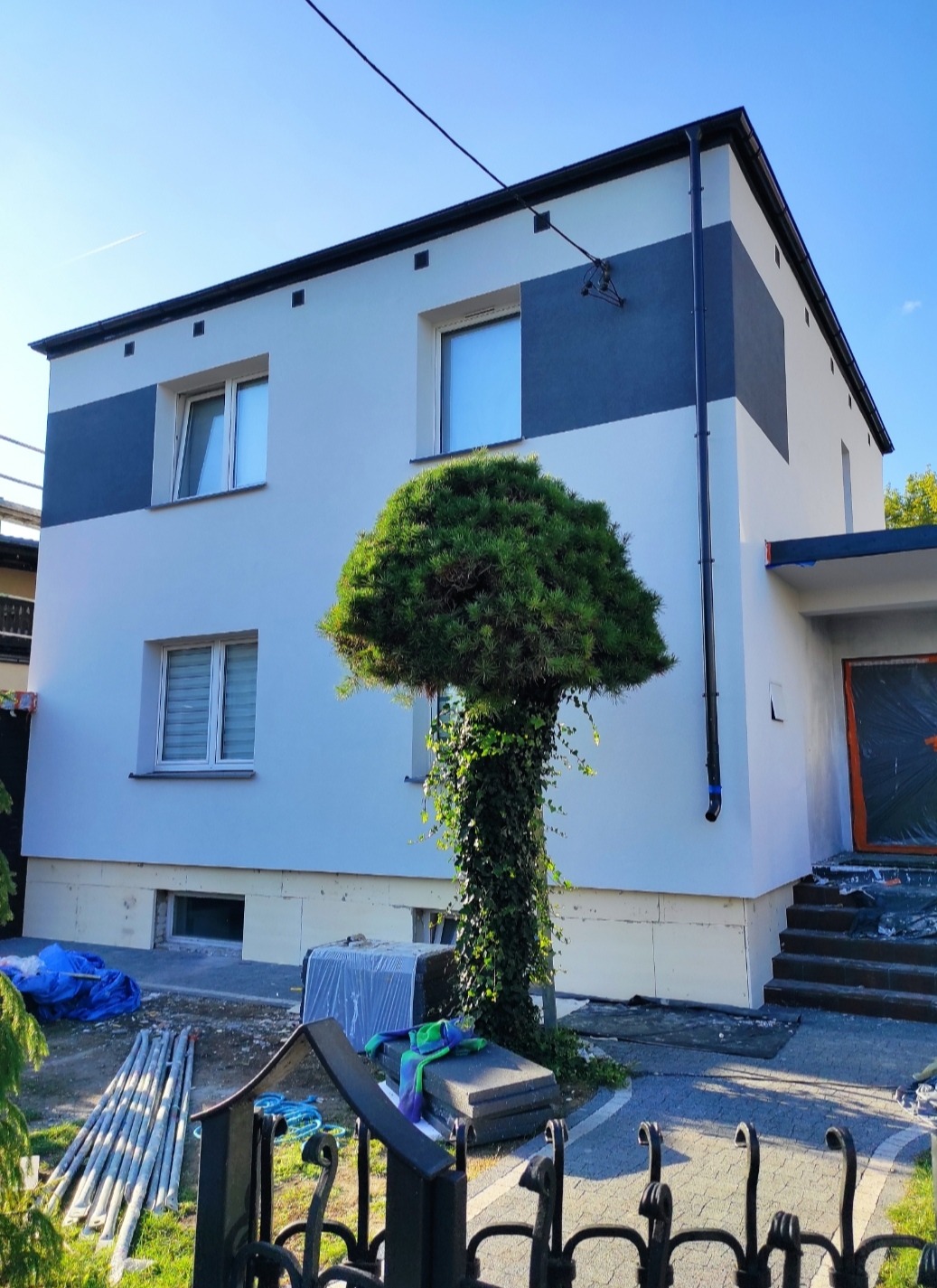 Dwukondygnacyjny dom po renowacji elewacji z szarym pasem, widoczne ocieplenie fundamentów i ozdobne drzewo przed wejściem. Błękitne niebo w tle.