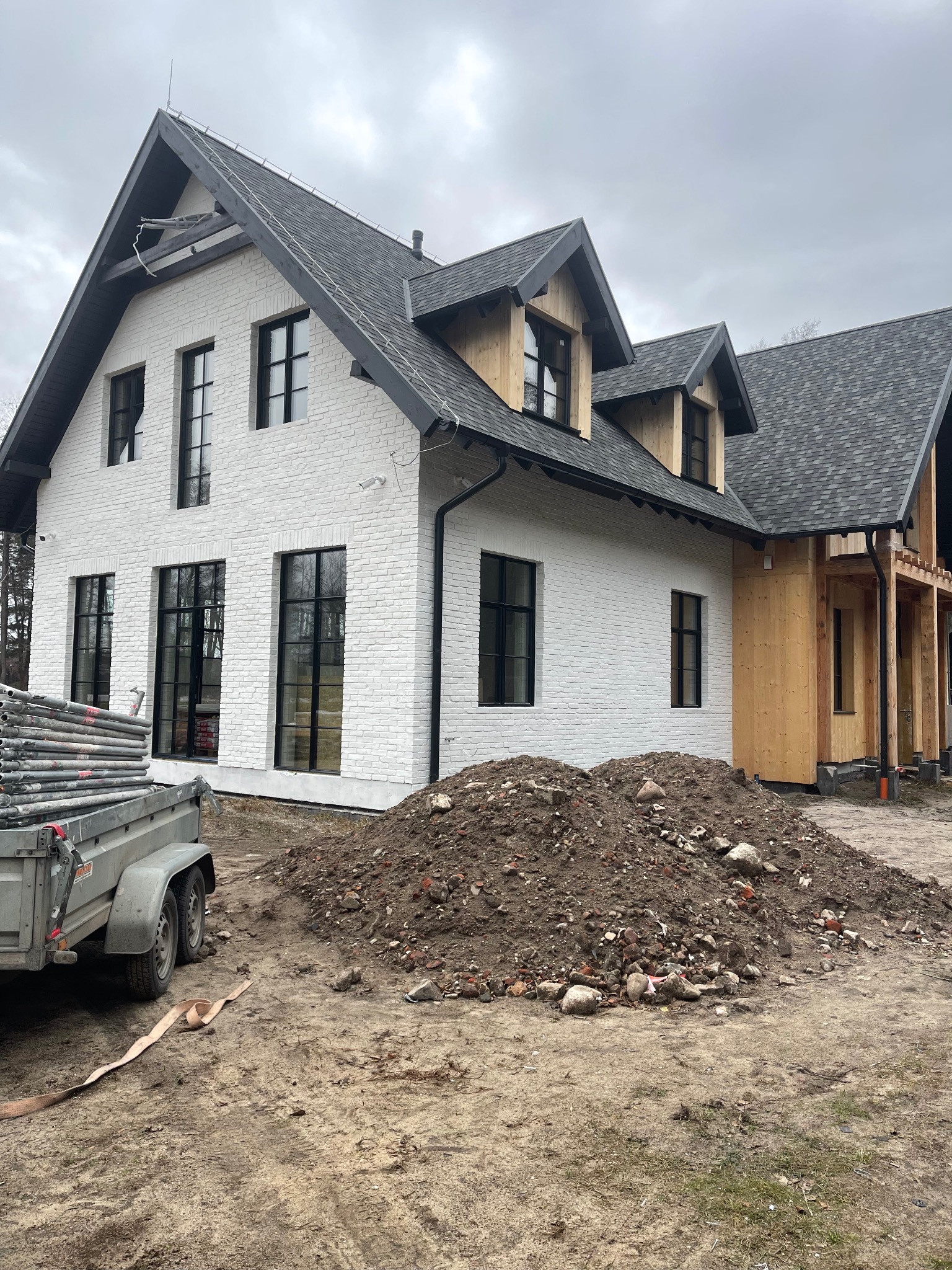 Dom w trakcie budowy: biała cegła, czarne okna, szary dach. Widoczny fragment drewnianej konstrukcji i przyczepa ze rusztowaniami. Na pierwszym planie hałda ziemi.