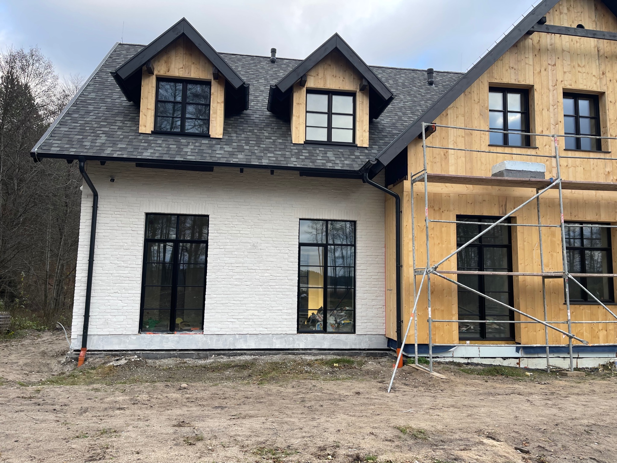 Dwukondygnacyjny dom z elewacją częściowo wykończoną białą cegłą i drewnem, z czarnymi ramami okiennymi, rusztowaniem i szarym dachem. Widok z zewnątrz.