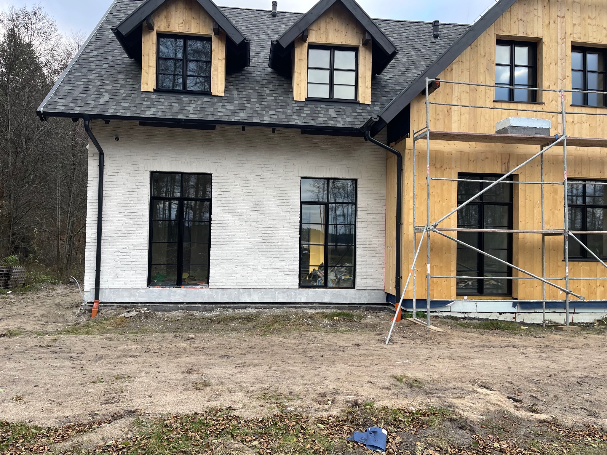 Dom w trakcie wykończenia elewacji: biała cegła i surowe drewno, czarne ramy okienne, rusztowanie. Widoczne detale wykończeniowe i etap prac.