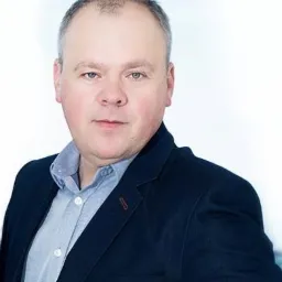 Portret mężczyzny w niebieskiej koszuli i granatowej marynarce, na jasnym tle.