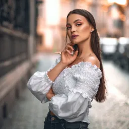 Portret młodej kobiety w białej bluzce z odkrytymi ramionami i ciemnych jeansach, pozującej na tle miejskiej uliczki z rozmytym tłem.