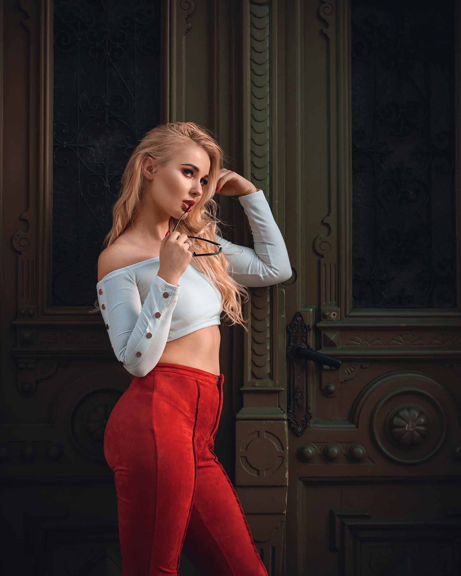 Portret kobiety o blond włosach w białym topie i czerwonych spodniach, pozującej na tle ozdobnych, ciemnozielonych drzwi