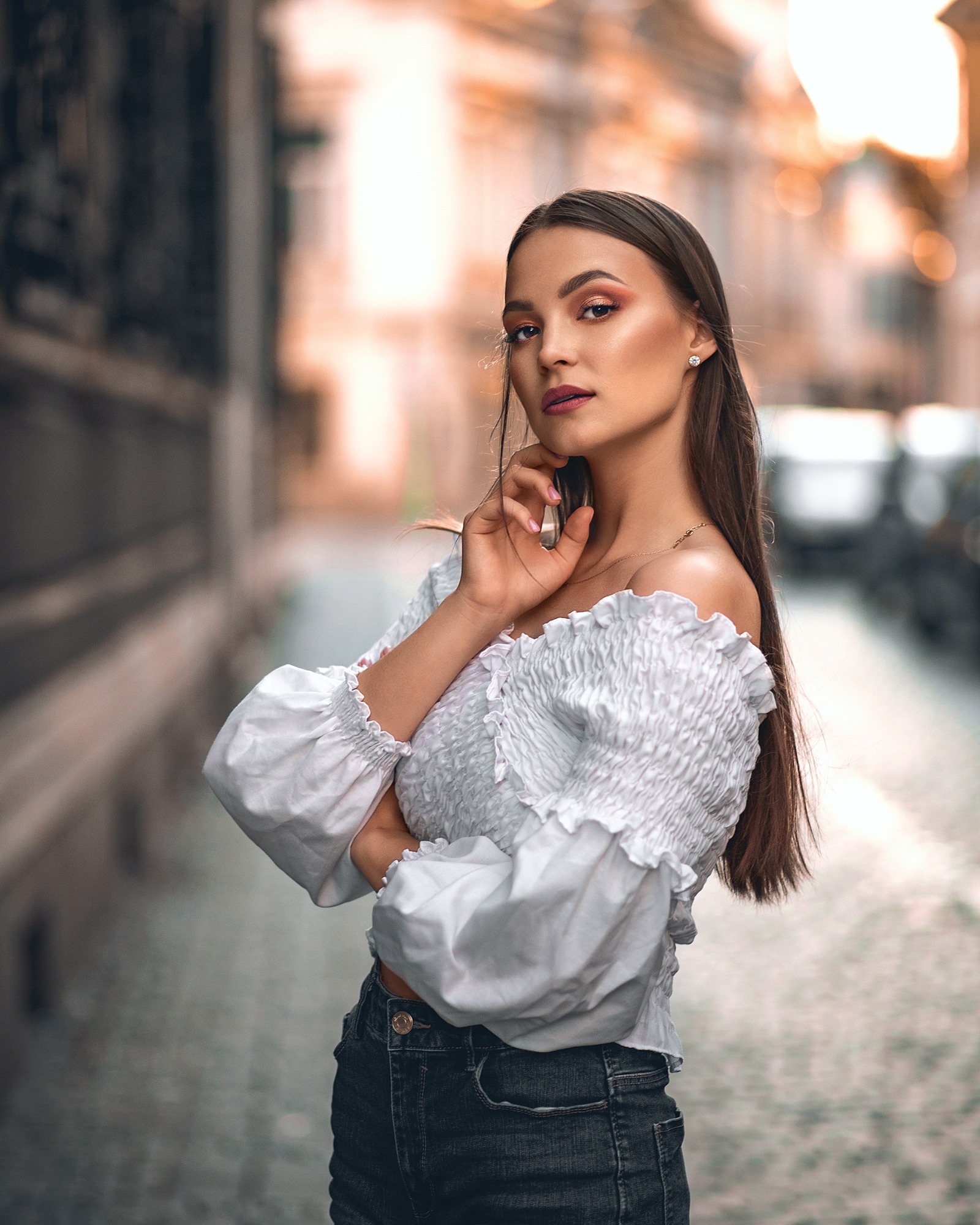 Portret młodej kobiety w białej bluzce z odkrytymi ramionami i ciemnych jeansach, pozującej na tle miejskiej uliczki z rozmytym tłem.