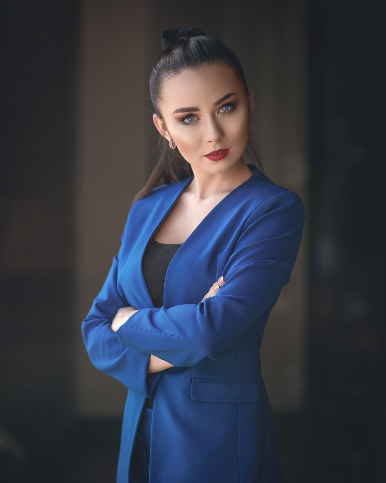 Portret kobiety w niebieskim garniturze, z założonymi rękami, patrzącej w obiektyw z wyrazistym makijażem i upiętymi włosami.