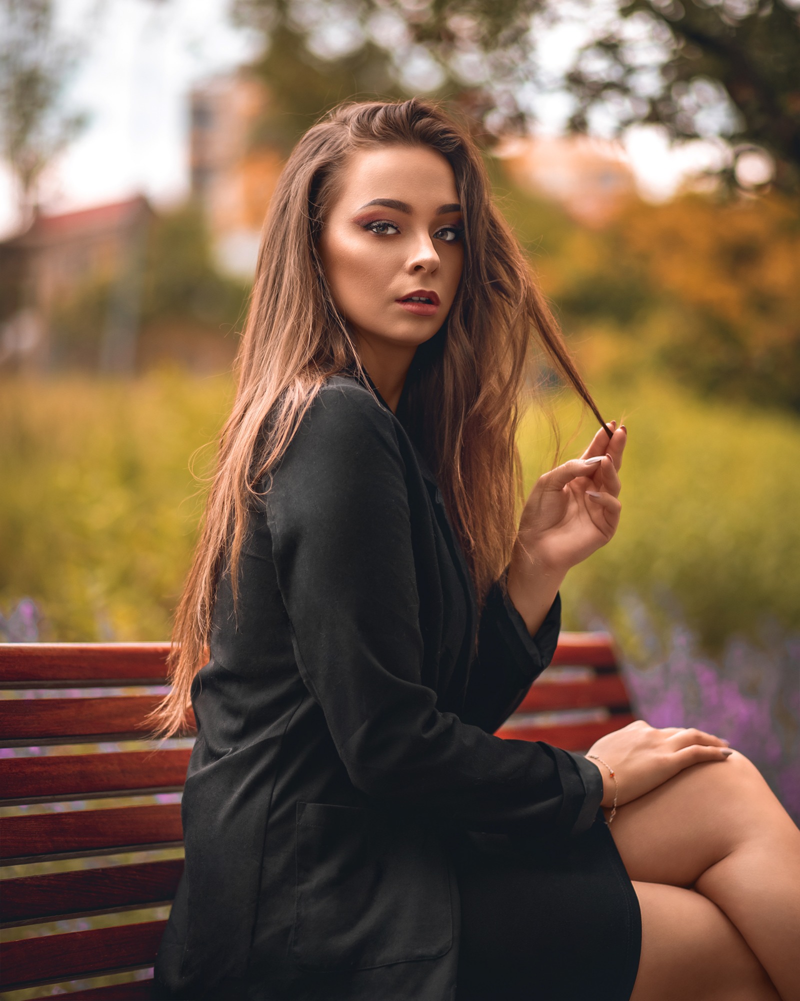 Portret kobiety w czarnej marynarce siedzącej na ławce w parku, trzymającej kosmyk włosów, z makijażem oczu w odcieniach różu i błyszczącymi ustami, w tle rozmyta zieleń i fioletowe kwiaty.