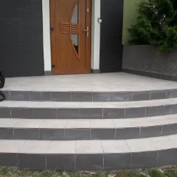 Wykończone schody wejściowe do domu, wyłożone jasnymi płytkami z ciemnoszarymi krawędziami, prowadzące do drewnianych drzwi wejściowych; czarna torba leży na pierwszym stopniu.