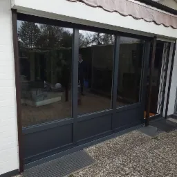 Ciemnoszare, trzyskrzydłowe drzwi tarasowe przesuwne z widocznym odbiciem osoby w szybie, zamontowane w białej, murowanej ścianie z zewnętrznym zadaszeniem.