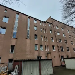 Fasada bloku mieszkalnego z licznymi oknami o białych ramach, widok z dołu, z widocznymi garażami blaszanymi i gałęziami drzew na pierwszym planie.