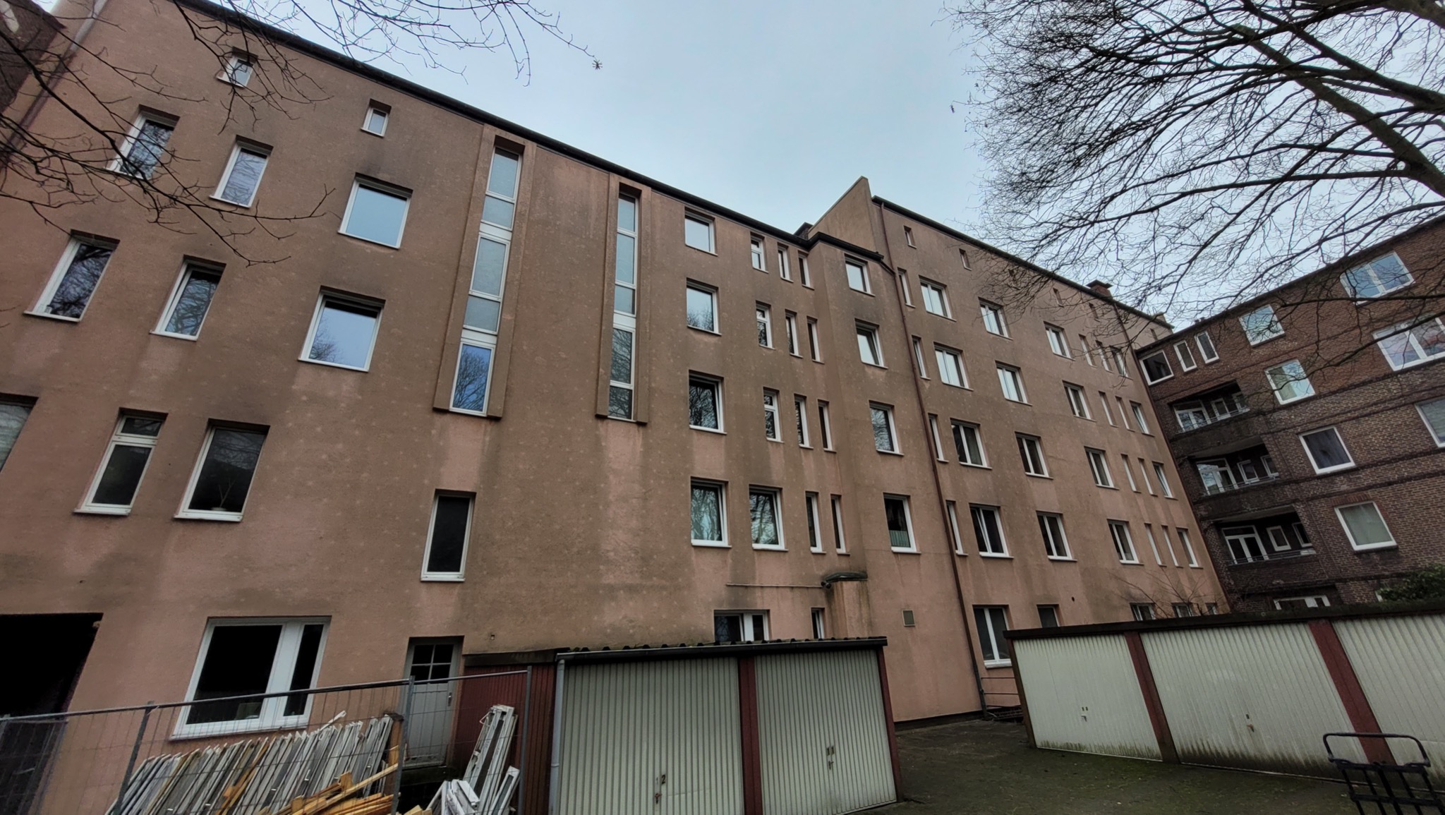 Fasada bloku mieszkalnego z licznymi oknami o białych ramach, widok z dołu, z widocznymi garażami blaszanymi i gałęziami drzew na pierwszym planie.