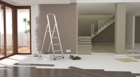 Wnętrze w trakcie remontu: drabina oparta o ścianę z częściowo pomalowaną powierzchnią, puszki z farbą, wałek malarski, panele podłogowe przygotowane do ułożenia, widok na schody w tle.
