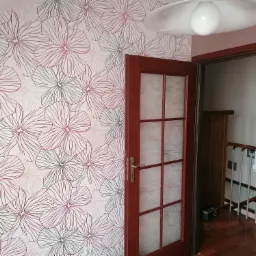 Ściana z tapetą w kwiaty w odcieniach szarości i różu, widoczna częściowo przeszklona, mahoniowa rama drzwi wewnętrznych, lampa sufitowa w tle.