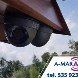 Dwie kamery monitoringu kopułkowe marki MS Vision zamontowane pod drewnianym okapem, widok z dołu na tle nieba i drzew, w prawym dolnym rogu logo firmy A-MARANT.