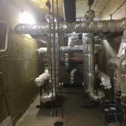 Izolowane rury w kotłowni z wykończonymi ścianami wełną mineralną, widoczne pompy i manometry, na podłodze otwór rewizyjny.