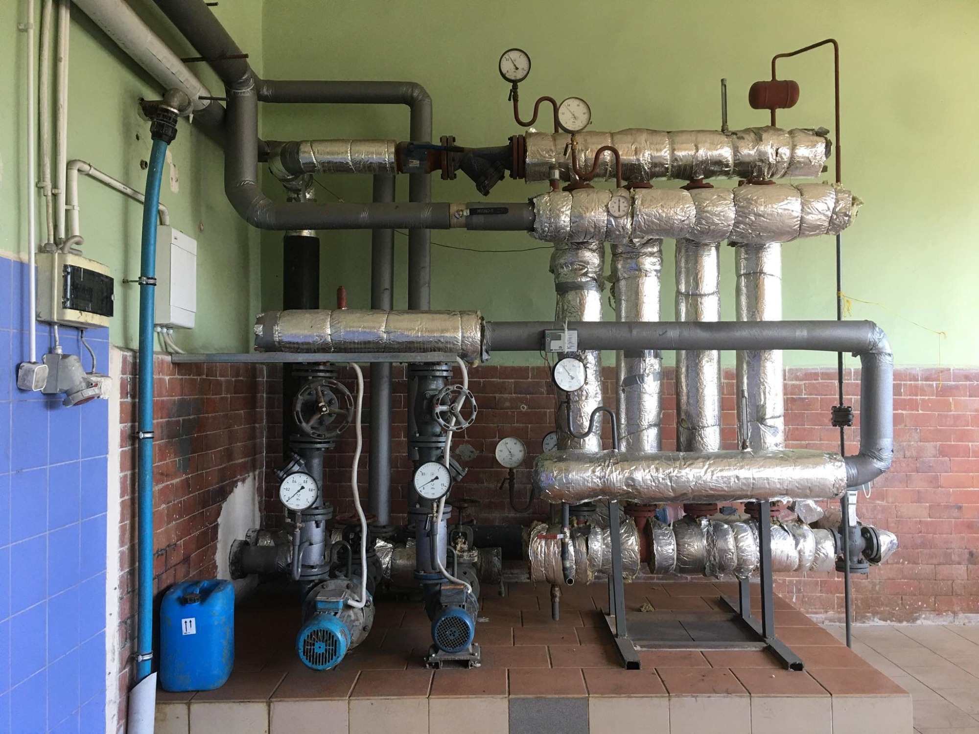 Fragment kotłowni z systemem rur i zaworów, izolowanych folią aluminiową, manometry wskazujące ciśnienie, niebieski kanister z płynem obok ceglanej ściany.
