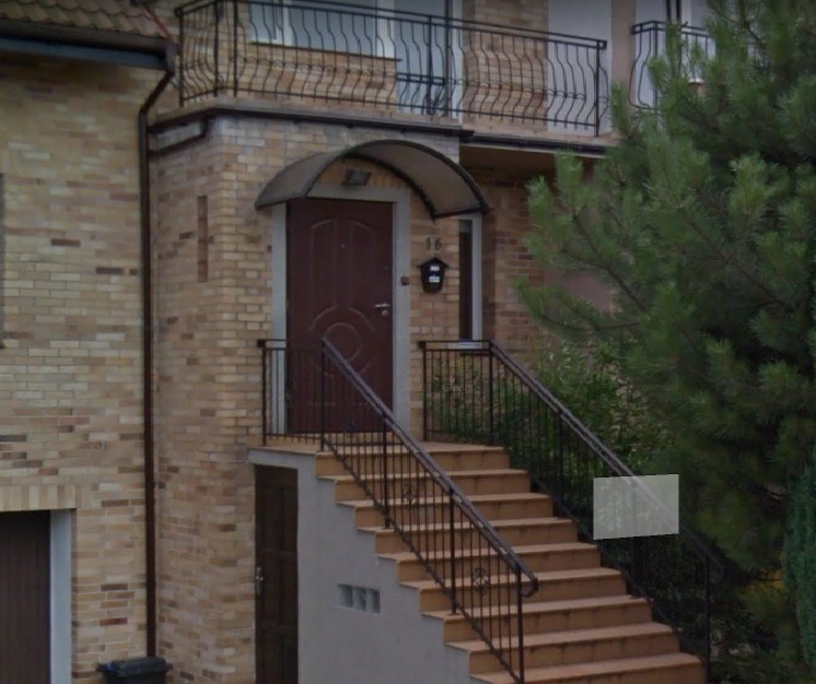 Wejście do budynku z brązowymi drzwiami, zadaszeniem i schodami z metalową barierką, na tle ściany z cegły klinkierowej i zieleni.