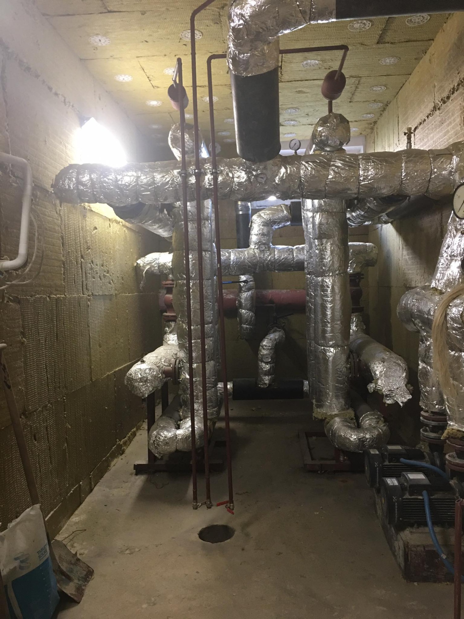 Izolowane rury w kotłowni z wykończonymi ścianami wełną mineralną, widoczne pompy i manometry, na podłodze otwór rewizyjny.