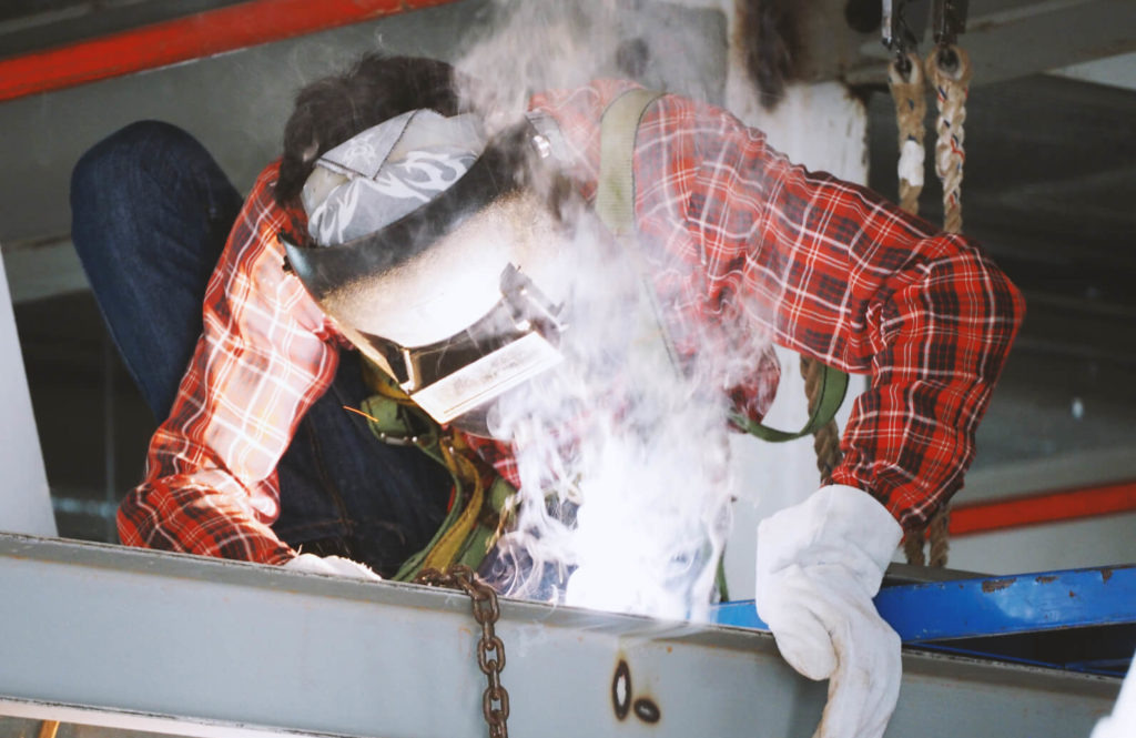 Osoba w masce ochronnej i flanelowej koszuli spawa metalową konstrukcję, widoczny dym i iskry, łańcuch w tle.