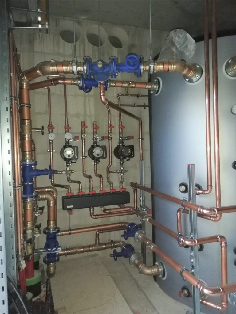 Złożony system rur miedzianych z niebieskimi zaworami, manometry i czujniki zamontowane na betonowej ścianie w pomieszczeniu technicznym, widoczny duży zbiornik.