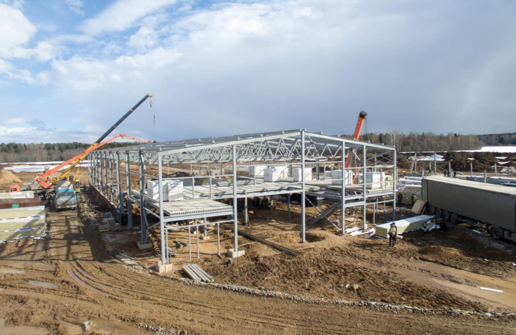 Wznoszenie stalowej konstrukcji hali przemysłowej z użyciem dźwigów na placu budowy, widoczne elementy konstrukcyjne i materiały budowlane.