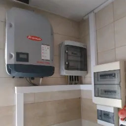 Zamontowany inwerter Fronius Symo na ścianie z beżowych płytek, obok skrzynki elektryczne z widocznymi przewodami, instalacja wewnątrz budynku.