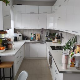 Paweł Pytlkowski - Nowoczesna, biała kuchnia w kształcie litery U z błyszczącymi frontami szafek, szarymi płytkami i drewnianymi akcentami. Na blacie widoczne akcesoria kuchenne i owoce.