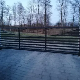 Przesuwna brama garażowa z poziomymi panelami, widok z zewnątrz na tle drzew bez liści i kostki brukowej.