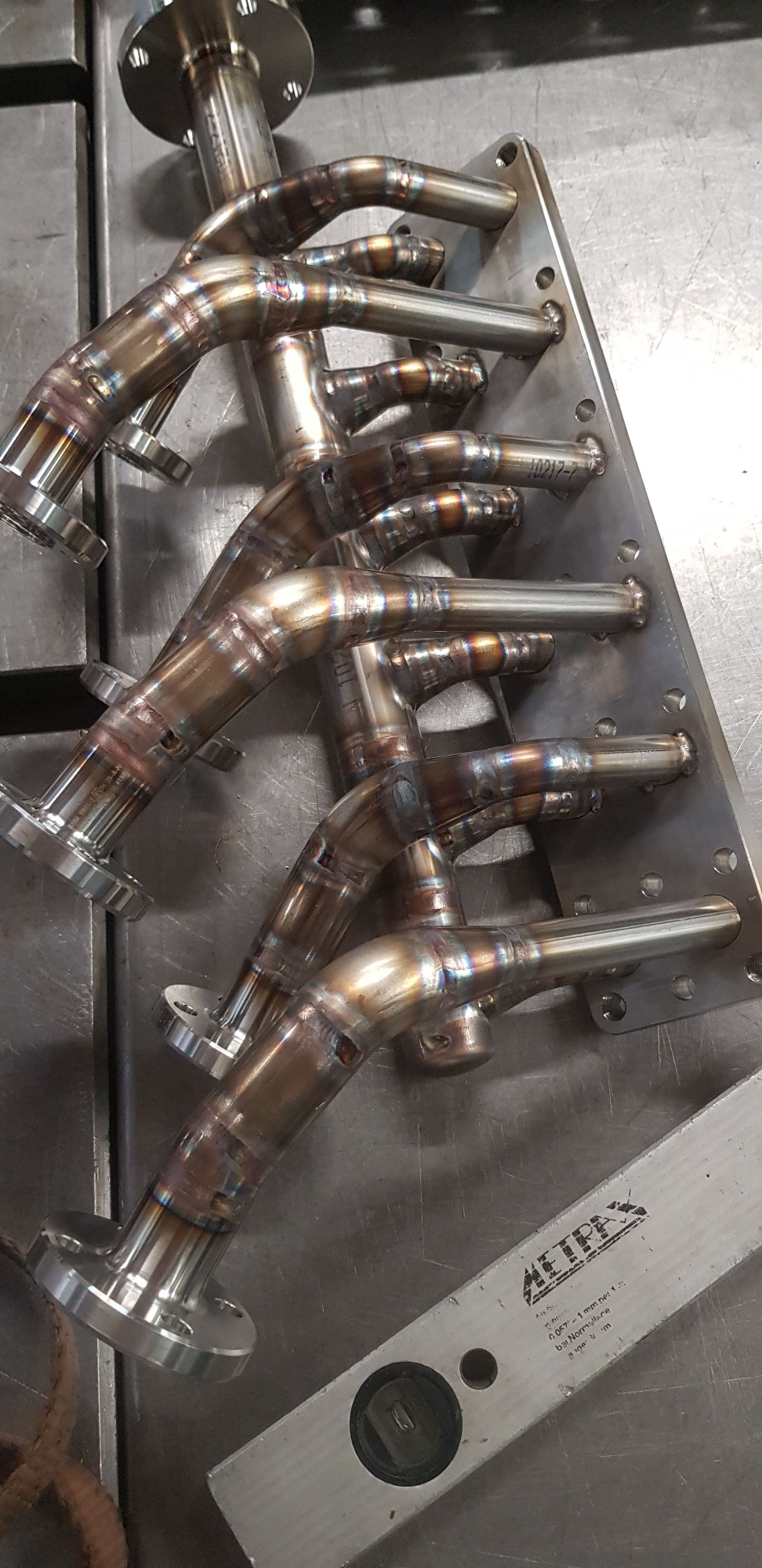 Zespawane kolektory rurowe z kołnierzami, ułożone na metalowym stole warsztatowym, widoczne ślady spawania i przebarwienia termiczne na metalu, obok poziomica.