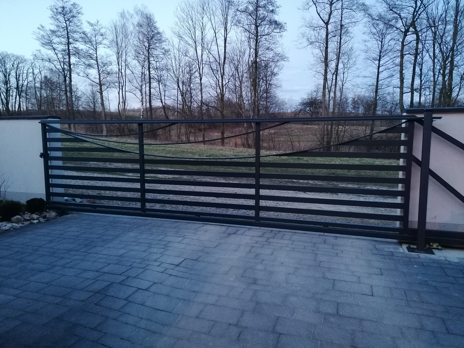 Przesuwna brama garażowa z poziomymi panelami, widok z zewnątrz na tle drzew bez liści i kostki brukowej.