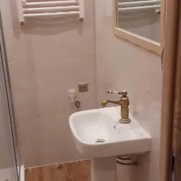 Biała umywalka na postumencie z mosiężną baterią w wyremontowanej łazience z brązowymi płytkami podłogowymi, grzejnik drabinkowy i lustro z dekoracyjnym ornamentem.