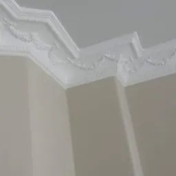 Dekoracyjny, biały gzyms sufitowy z ornamentami, zamontowany na styku ściany i sufitu w jasnym pomieszczeniu.