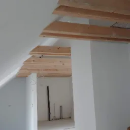 Surowe drewniane belki stropowe na poddaszu w trakcie prac w Sikuty, widoczne przewody elektryczne i fragmenty instalacji.