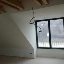 Poddasze w trakcie remontu: drewniane belki stropowe, ściany po gładziowaniu, wystające kable elektryczne, okno z widokiem na dach sąsiedniego budynku.