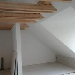 Poddasze w trakcie prac wykończeniowych: widoczne drewniane belki stropowe, białe ściany ze skośnym sufitem, płyty gipsowo-kartonowe oparte o ścianę, przygotowane do montażu.