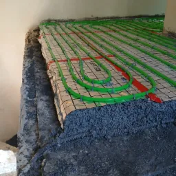 Montaż ogrzewania podłogowego: zielone rury grzewcze ułożone na folii z siatką, przymocowane plastikowymi uchwytami, zalane warstwą wylewki z widoczną rurą karbowaną w podłodze.