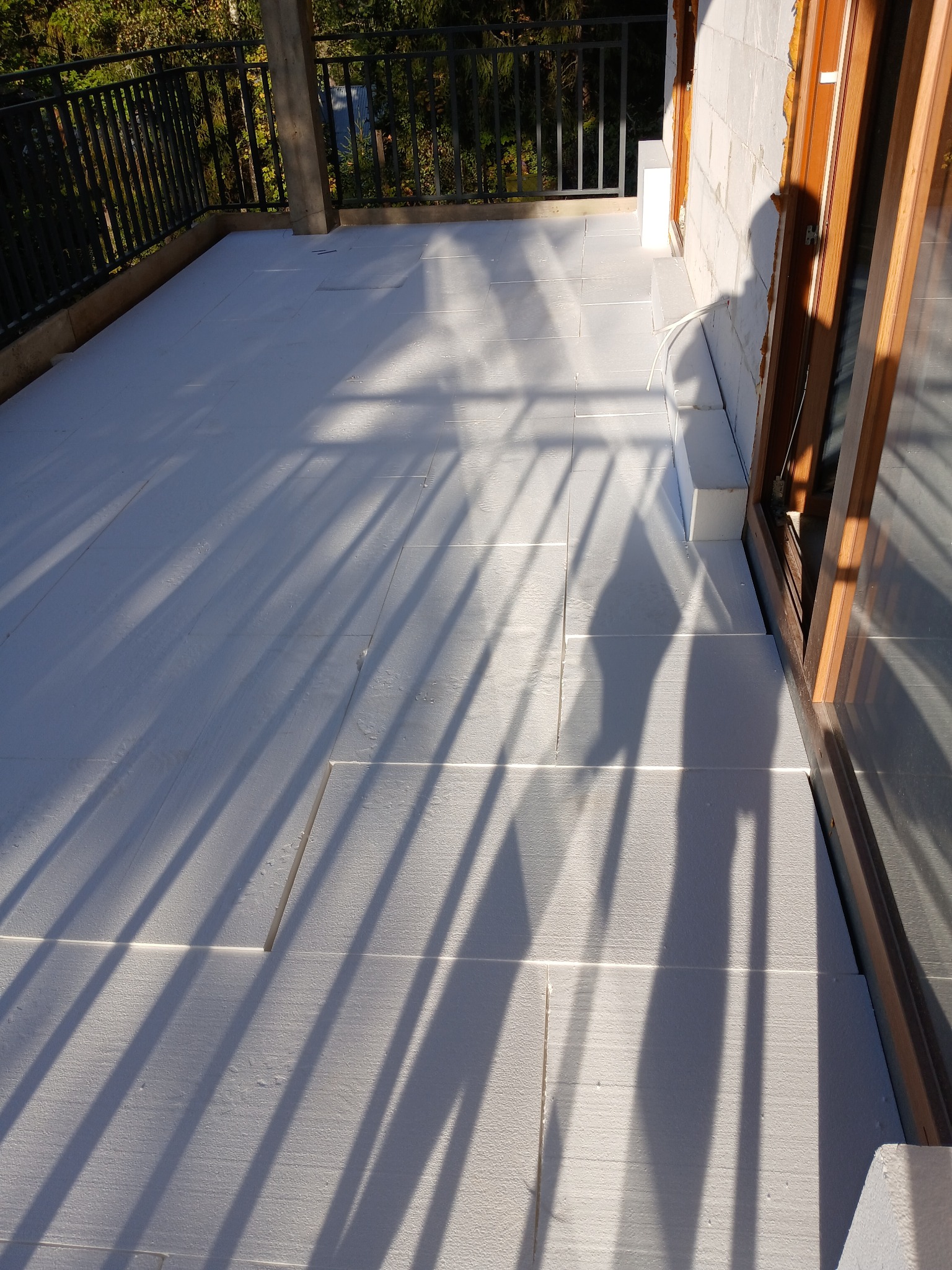 Białe płyty izolacyjne ułożone na balkonie, widoczne cienie barierki i fragment okna z drewnianą ramą.