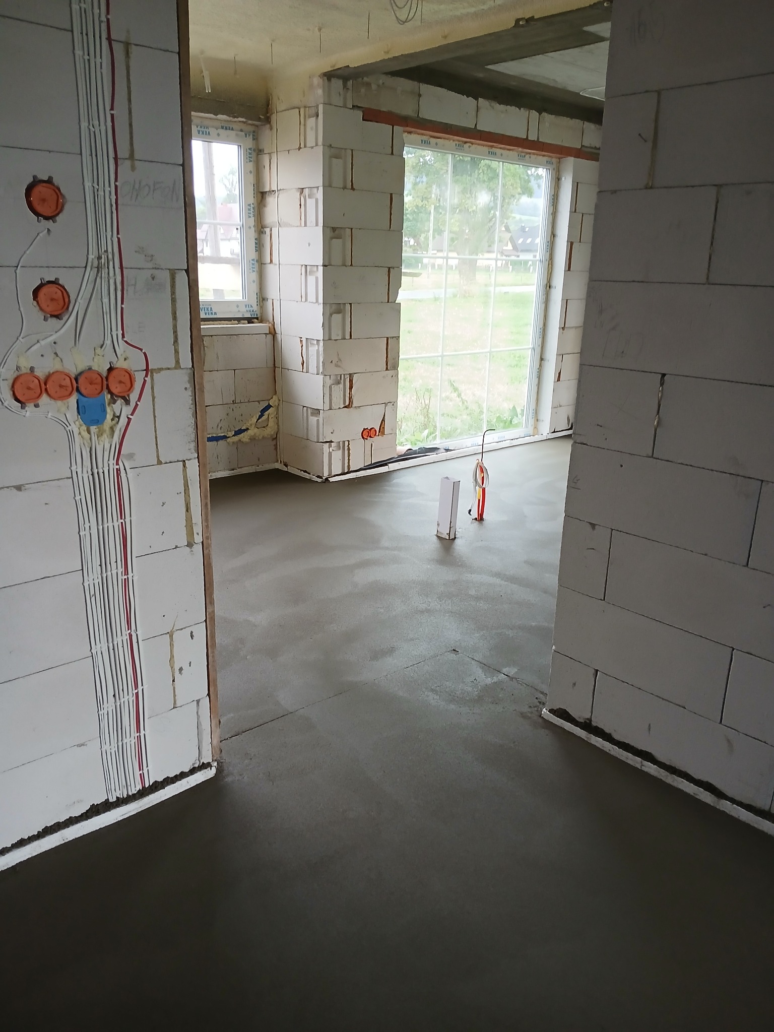 Świeżo wylana, gładka posadzka w surowym wnętrzu budynku w trakcie budowy, widoczne okna, instalacja elektryczna w ścianie oraz białe bloczki konstrukcyjne.