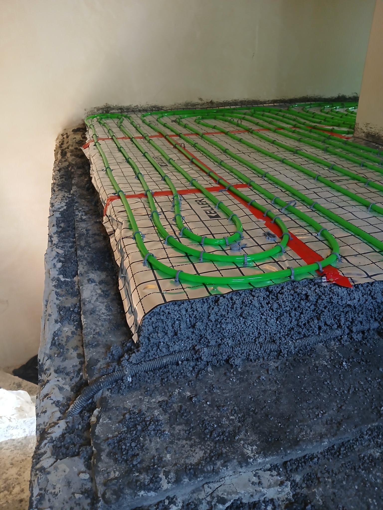 Montaż ogrzewania podłogowego: zielone rury grzewcze ułożone na folii z siatką, przymocowane plastikowymi uchwytami, zalane warstwą wylewki z widoczną rurą karbowaną w podłodze.